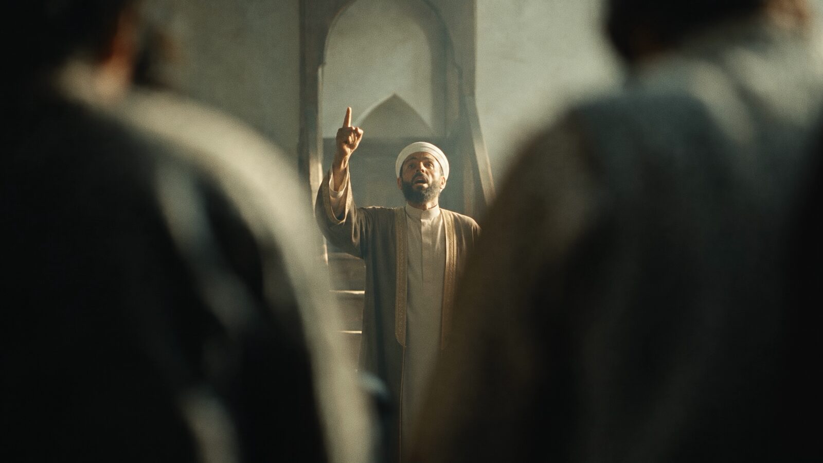 An imam raising a finger, seen past two shoulders.